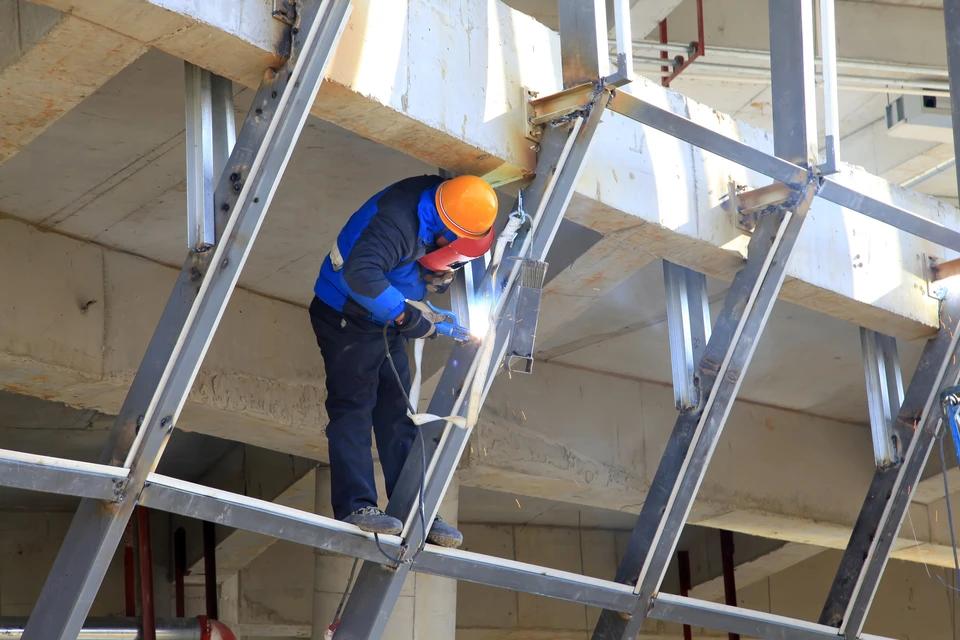 Chantier de construction intérieur, ouvrier en combinaison bleue et casque orange effectuant des travaux de soudure sur échafaudage métallique