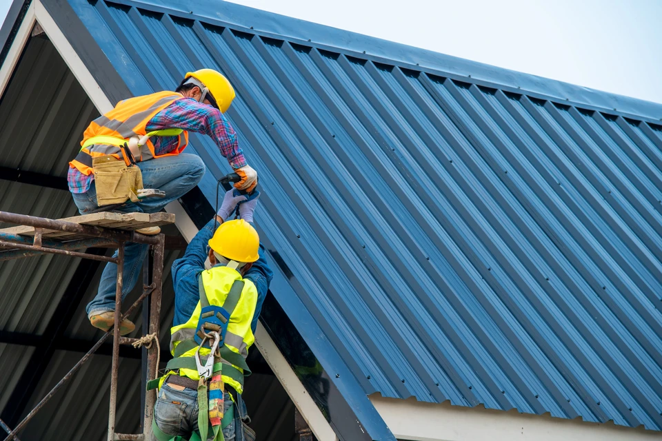 Deux ouvriers en équipement de sécurité (casques jaunes, gilets haute visibilité) installent une toiture en tôle ondulée bleue sur échafaudage